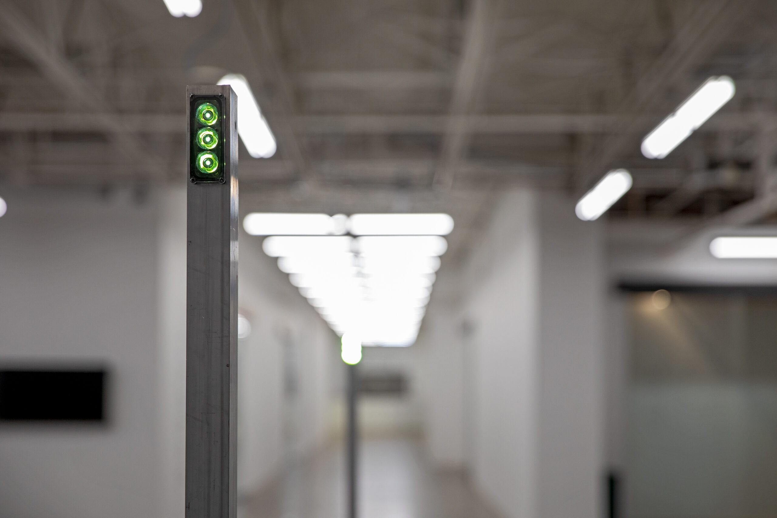Detailed view of a light rod's lime green LED lamp at low power in a hallway with blurred fluorescent lights receding into the distance